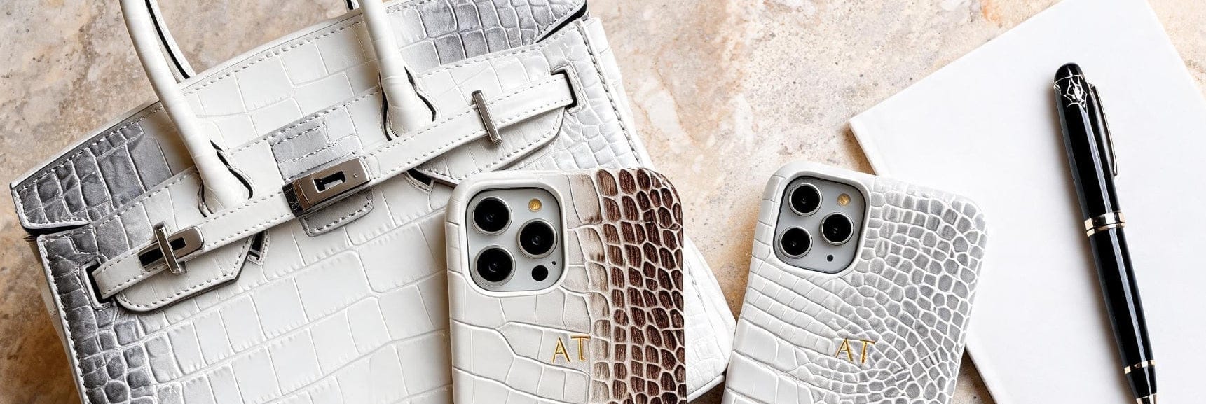 Two crocodile-patterned phone cases on a marble surface with a handbag and pen.